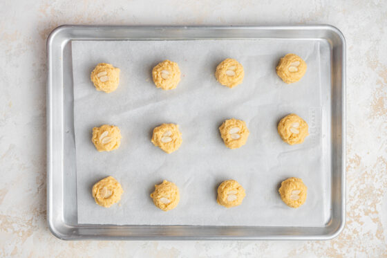 Almond cookie dough shaped into 12 balls on a parchment lined baking sheet topped with almond slivers.
