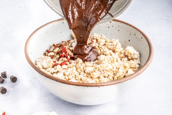 Pouring chocolate peanut butter mixture over rice cakes and mix-ins in a bowl.