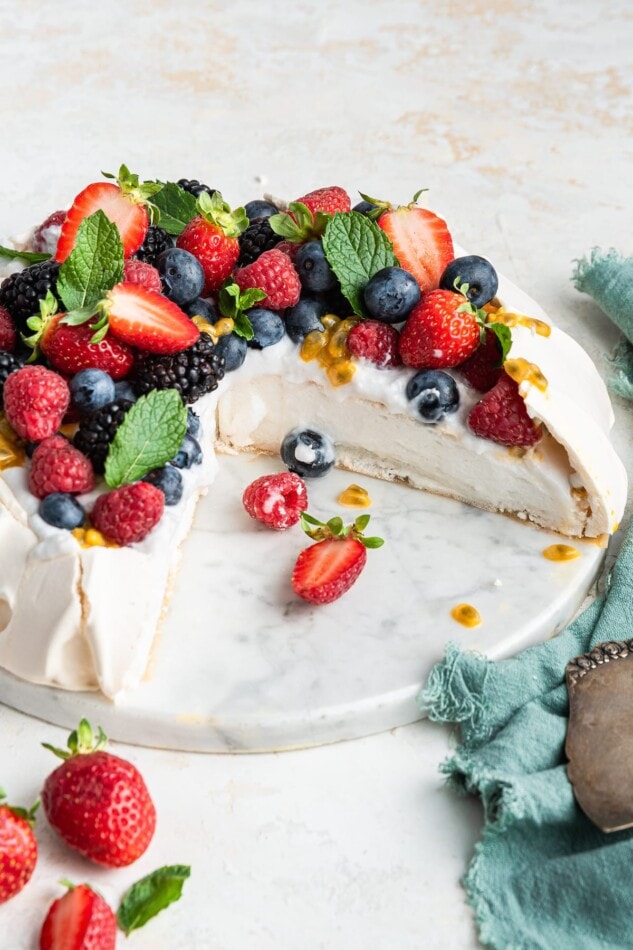 A pavlova topped with coconut whipped cream and fresh fruit with slices missing.