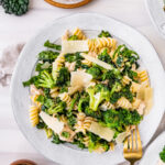 A large plate with a serving of pasta, kale, broccoli, tuna, and shaved parmesan.