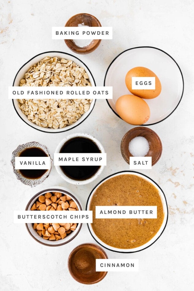 Ingredients measured out to make Oatmeal Scotchies: baking powder, oats, eggs, vanilla, maple syrup, salt, butterscotch chips, almond butter and cinnamon.