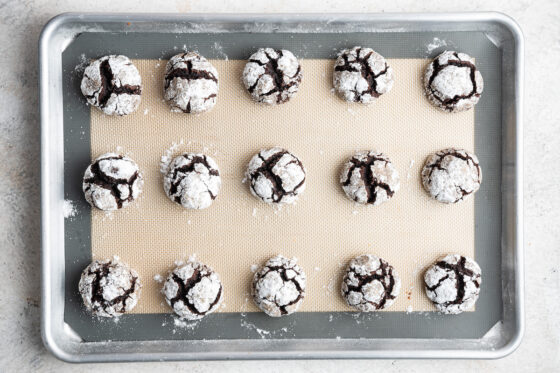 Fifteen baked chocolate crinkle cookies on a baking sheet lined with a silpat mat.