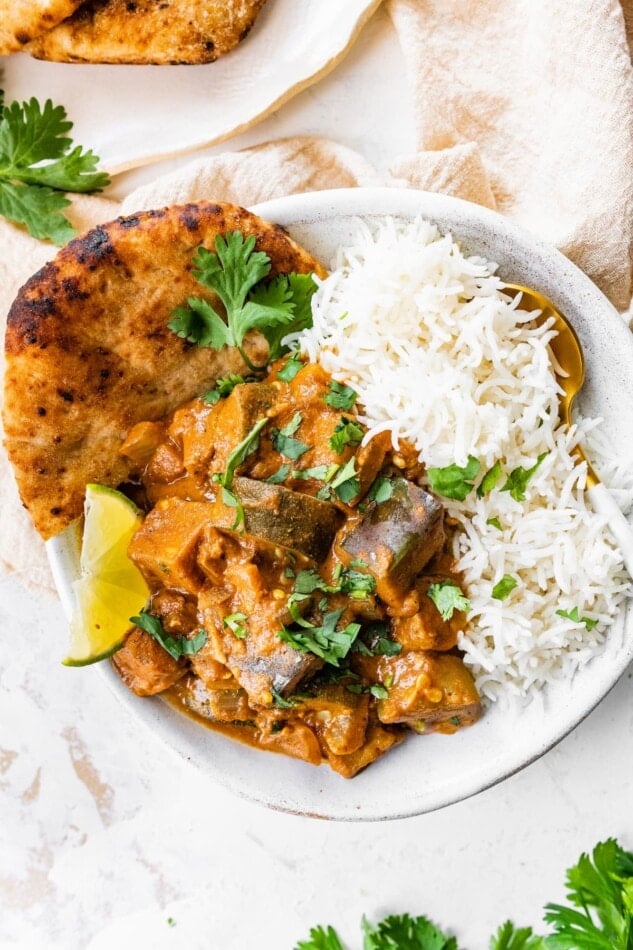 A shallow bowl of eggplant curry with rice and naan.