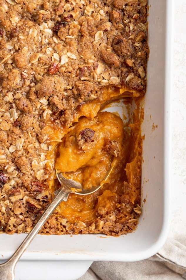 A serving spoon resting in a baking dish containing healthy sweet potato casserole.