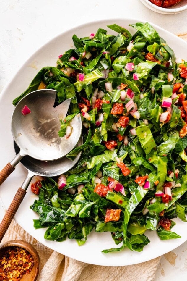 A serving bowl containing raw collard greens salad with salad servers.