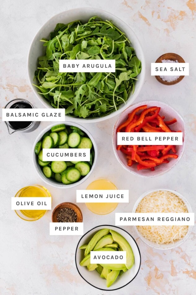 Ingredients measured out to make Bella Hadid Salad: baby arugula, sea salt, balsamic glaze, cucumbers, red bell pepper, olive oil, lemon juice, pepper, parmesan reggiano and avocado.