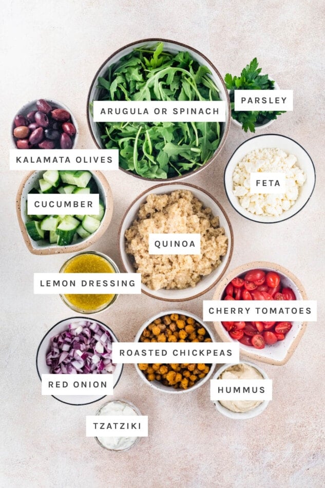Ingredients measured out to make Mediterranean Quinoa Bowl: arugula, parsley, kalamata olives, cucumber, feta, quinoa, lemon dressing, cherry tomatoes, roasted chickpeas, red onion, tzatziki and hummus.