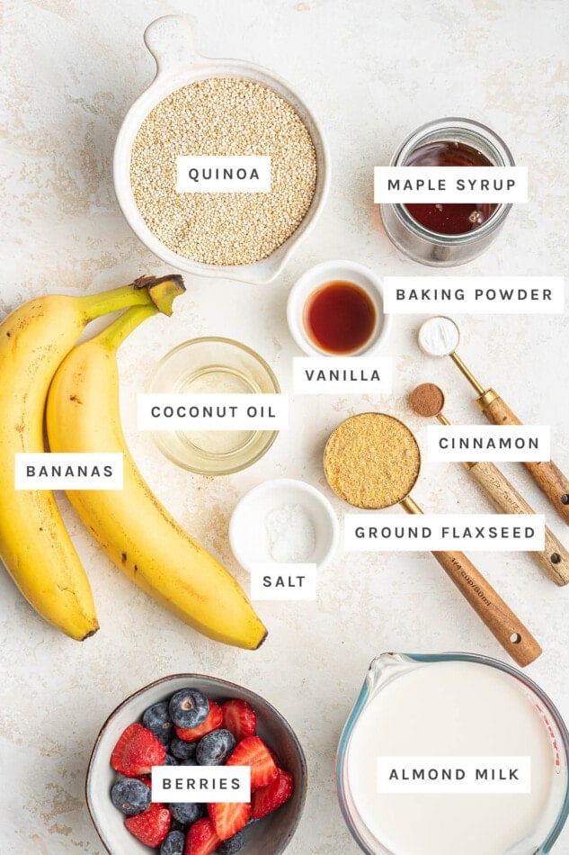 Ingredients measured out to make Quinoa Breakfast Bake: quinoa, maple syrup, baking powder, vanilla, coconut oil, cinnamon, bananas, ground flaxseed, salt, berries and almond milk.