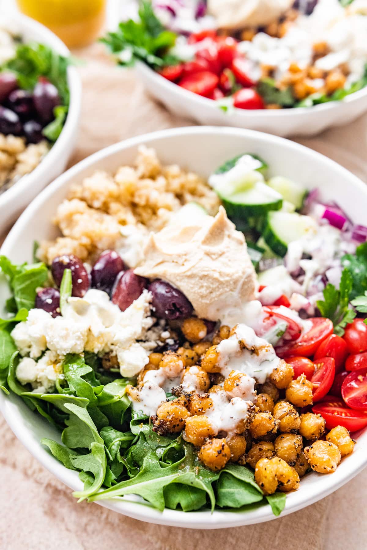 A Mediterranean Quinoa Bowl with quinoa, arugula, chickpeas, tomatoes, red onion, cucumbers, kalamata olives, feta cheese and hummus drizzled with tzatziki.