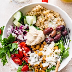 A Mediterranean Quinoa Bowl with quinoa, arugula, chickpeas, tomatoes, red onion, cucumbers, kalamata olives, feta cheese and hummus drizzled with tzatziki.