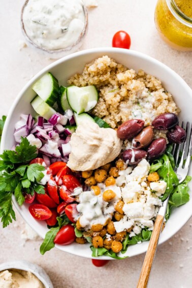 A Mediterranean Quinoa Bowl with quinoa, arugula, chickpeas, tomatoes, red onion, cucumbers, kalamata olives, feta cheese and hummus drizzled with tzatziki.