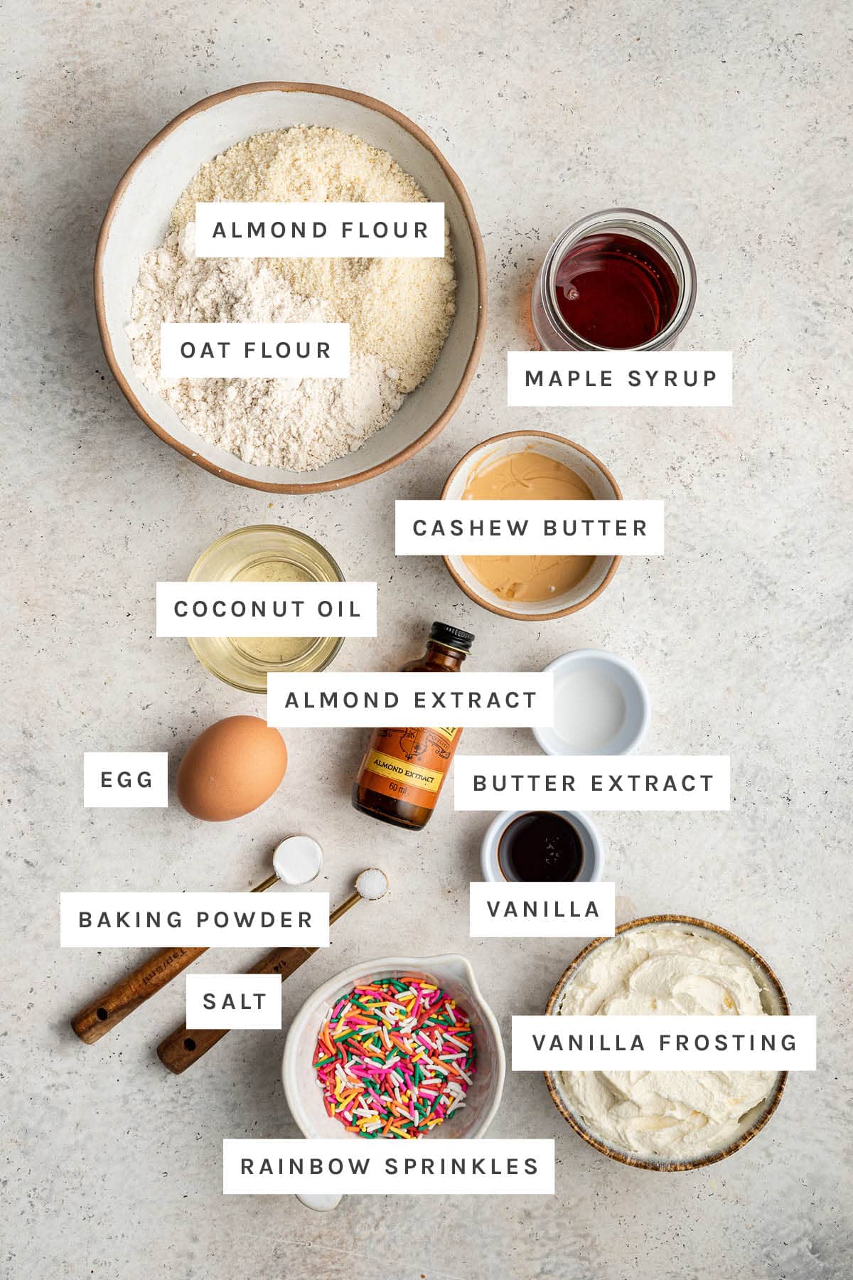 Ingredients measured out to make Birthday Cake Cookies: almond flour, oat flour, maple syrup, cashew butter, coconut oil, almond extract, butter extract, egg, baking powder, salt, vanilla, vanilla frosting and rainbow sprinkles.