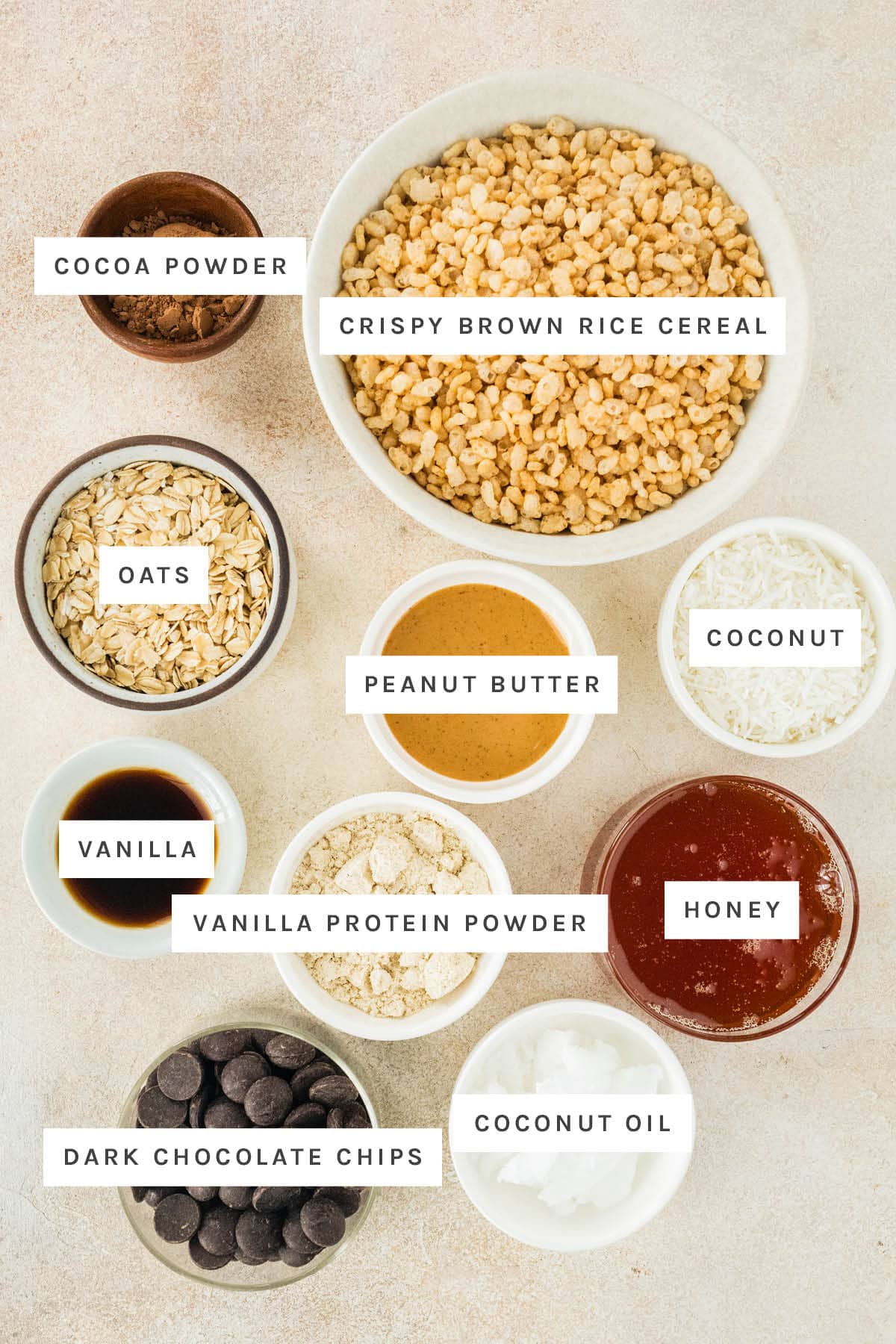 Ingredients measured out to make Crunchy Protein Bars: cocoa powder, crispy brown rice cereal, oats, peanut butter, coconut, vanilla, vanilla protein powder, honey, dark chocolate chips and coconut oil.