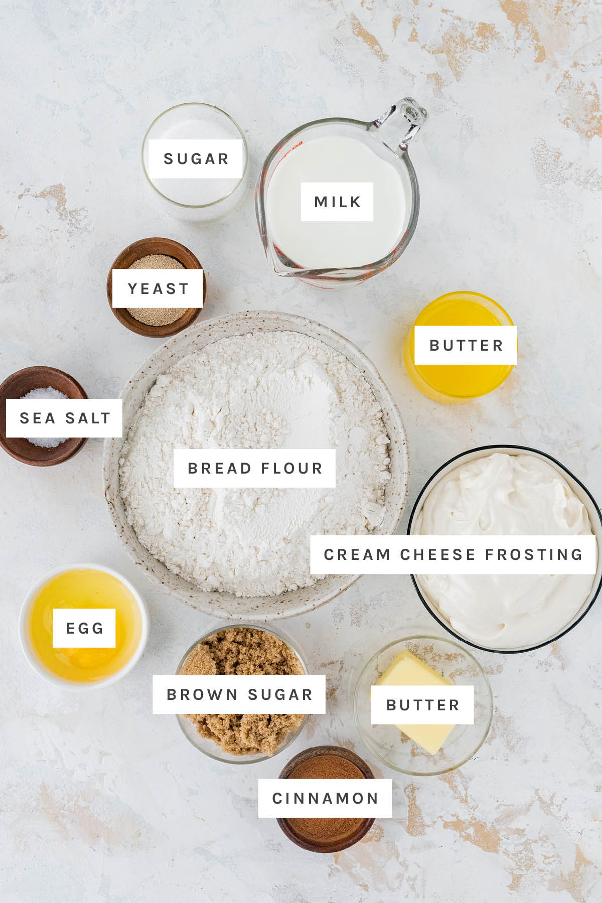 Ingredients measured out to make Fluffy Cinnamon Rolls: sugar, milk, yeast, butter, sea salt, bread flour, cream cheese frosting, egg, brown sugar and cinnamon.