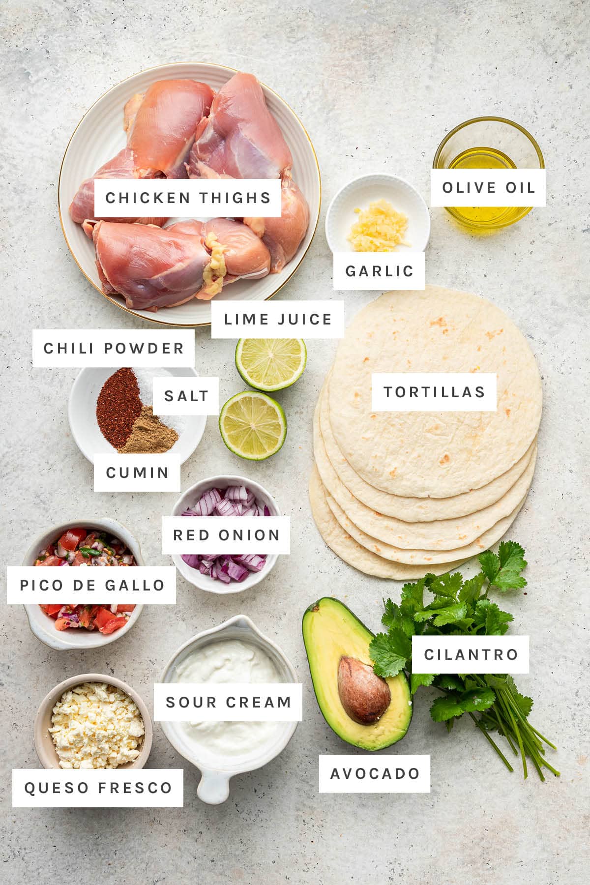 Ingredients measured out to make Healthy Chicken Tacos: chicken thighs, garlic, olive oil, lime, chili powder, salt, cumin, red onion, tortillas, pico de gallo, queso fresco, sour cream, avocado and cilantro.