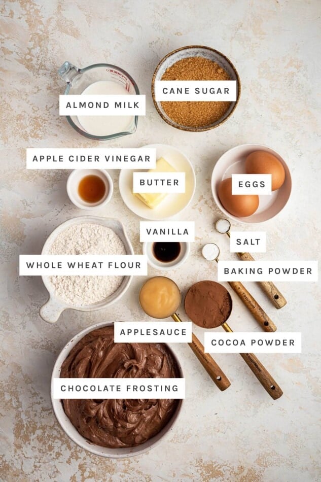 Ingredients measured out to make Healthy Chocolate Cupcakes: almond milk, cane sugar, apple cider vinegar, butter, eggs, vanilla, whole wheat flour, salt, baking powder, applesauce, cocoa powder and chocolate frosting.