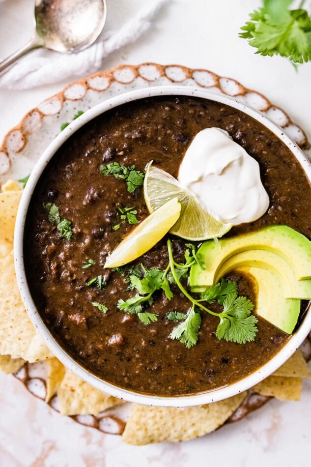 A bowl of black bean soup topped with avocado, cilantro, lime and vegan sour cream served alongside tortilla chips.