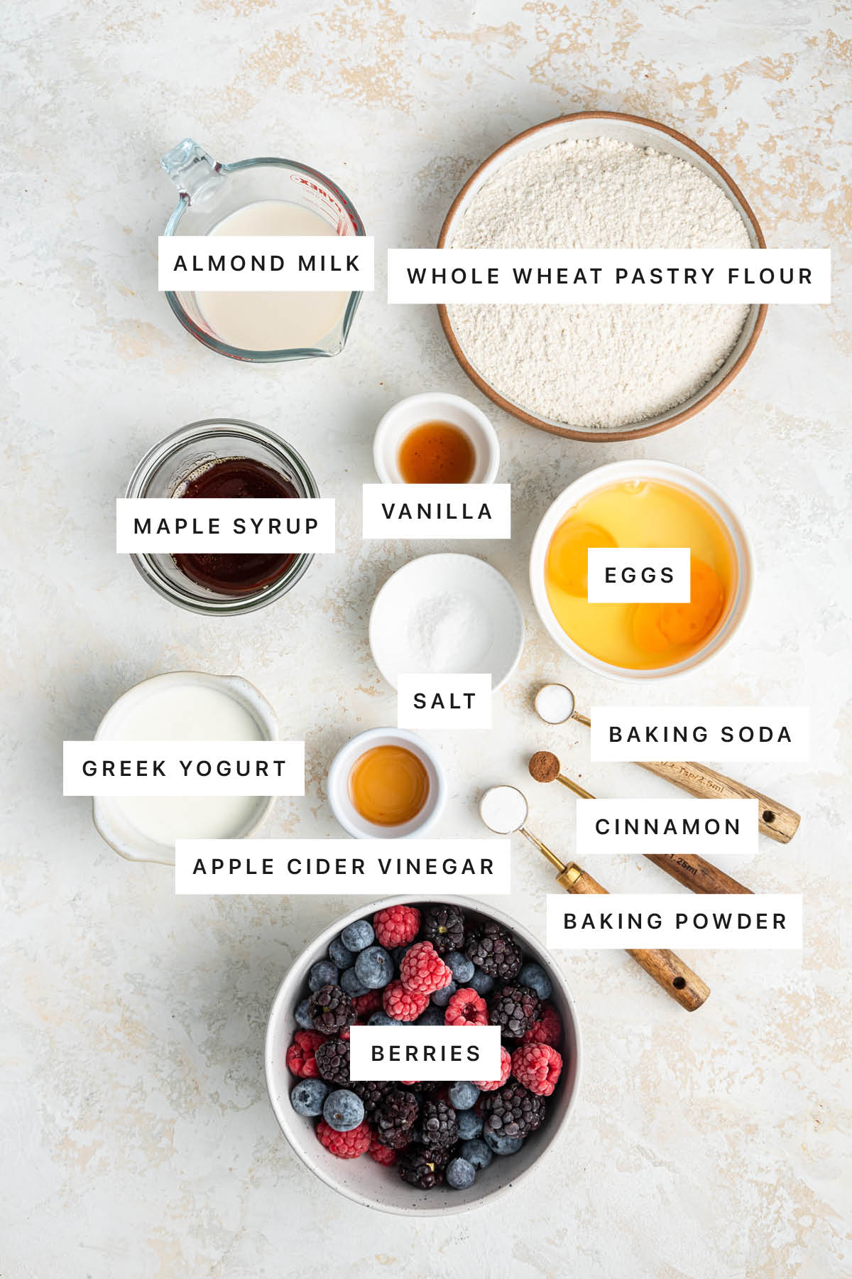 Ingredients measured out to make Easy Berry Cake: almond milk, whole wheat pastry flour, maple syrup, vanilla, eggs, salt, Greek yogurt, apple cider vinegar, baking soda, cinnamon, baking powder and berries.