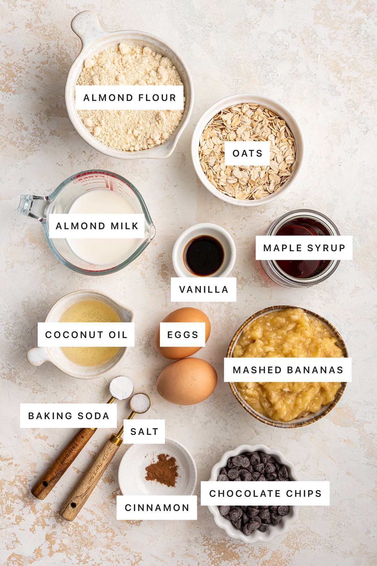 Ingredients measured out to make Banana Blender Muffins: almond flour, oats, almond milk, vanilla, maple syrup, coconut oil, eggs, mashed bananas, baking soda, salt, cinnamon and chocolate chips.