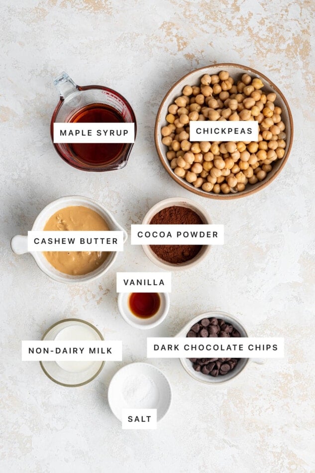 Ingredients measured out to make Chickpea Brownies: maple syrup, chickpeas, cashew butter, cocoa powder, vanilla, non-dairy milk, dark chocolate chips and salt.
