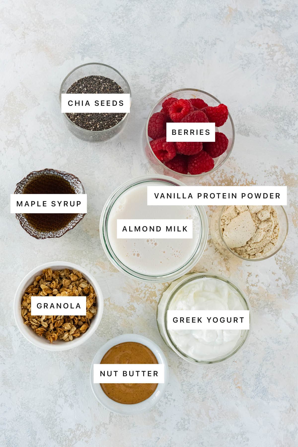 Ingredients measured out to make Protein Chia Pudding: chia seeds, berries, maple syrup, vanilla protein powder, almond milk, granola, Greek yogurt and nut butter.