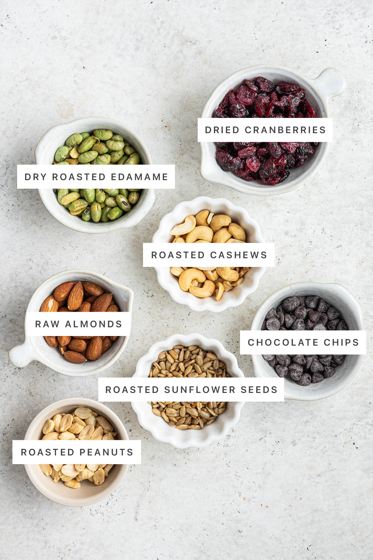 Ingredients measured out to make Protein Trail Mix: dry roasted edamame, dried cranberries, roasted cashews, raw almonds, chocolate chips, roasted sunflower seeds, roasted peanuts.