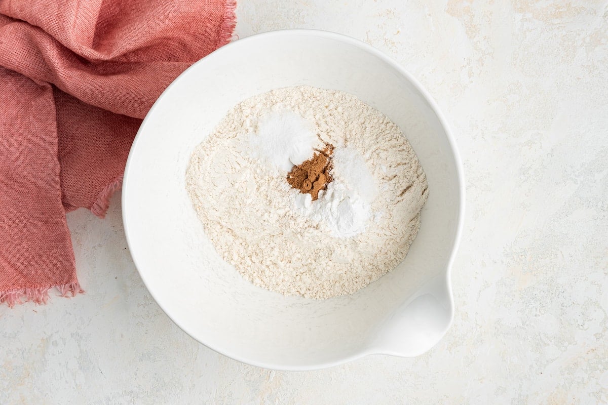 Flour, baking powder, baking soda, salt and cinnamon in a white medium mixing bowl.