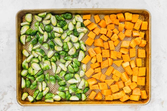 A sheet pan half covered with seasoned brussels sprouts and half covered in butternut squash chunks.