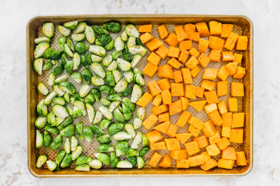 A sheet pan half covered with seasoned brussels sprouts and half covered in butternut squash chunks.