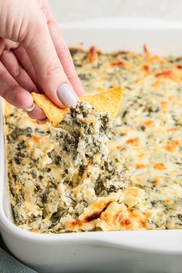 A hand dipping a chip into spinach artichoke dip.