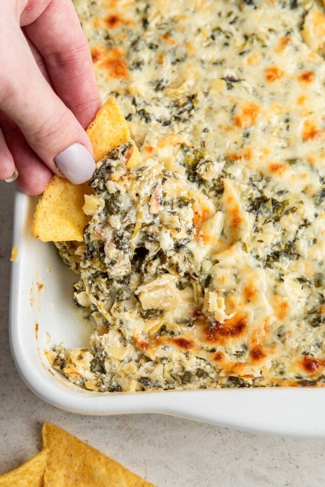 A hand dipping a chip into a dish of spinach artichoke dip.