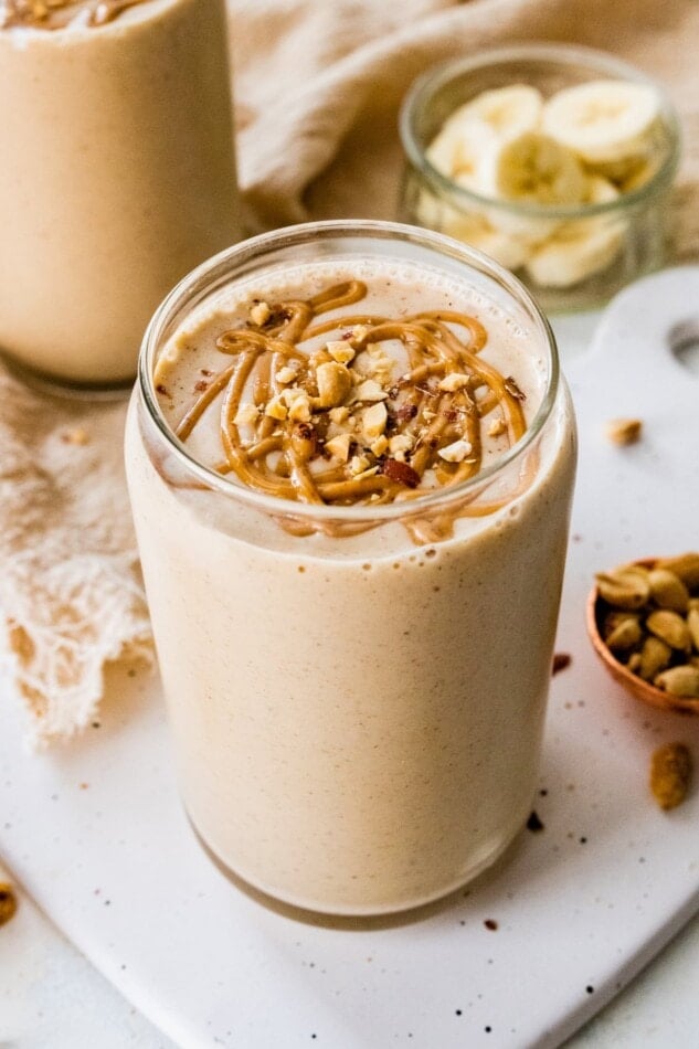Close up of a peanut butter protein shake in a glass with a drizzle of peanut butter and chopped peanuts on top.