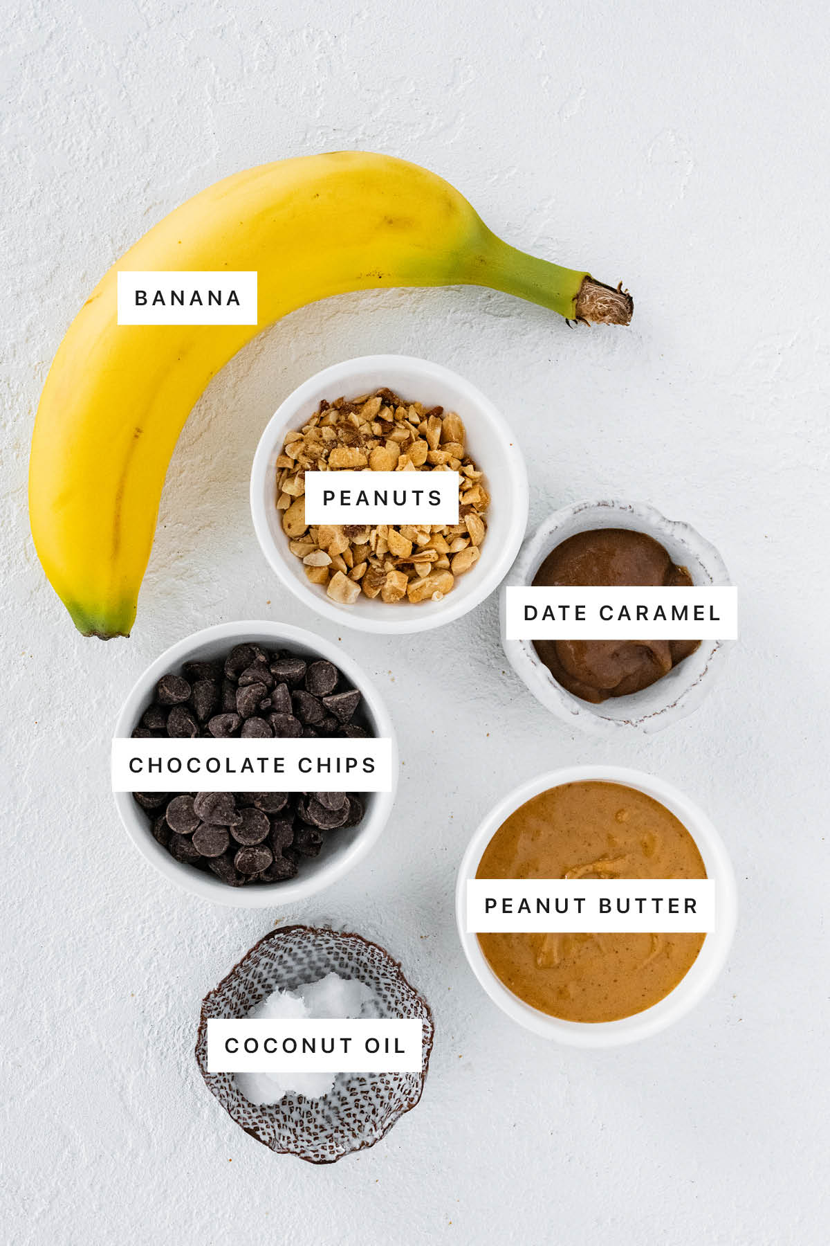 Ingredients measured out to make Frozen Banana Snickers: banana, peanuts, date caramel, chocolate chips, peanut butter and coconut oil.
