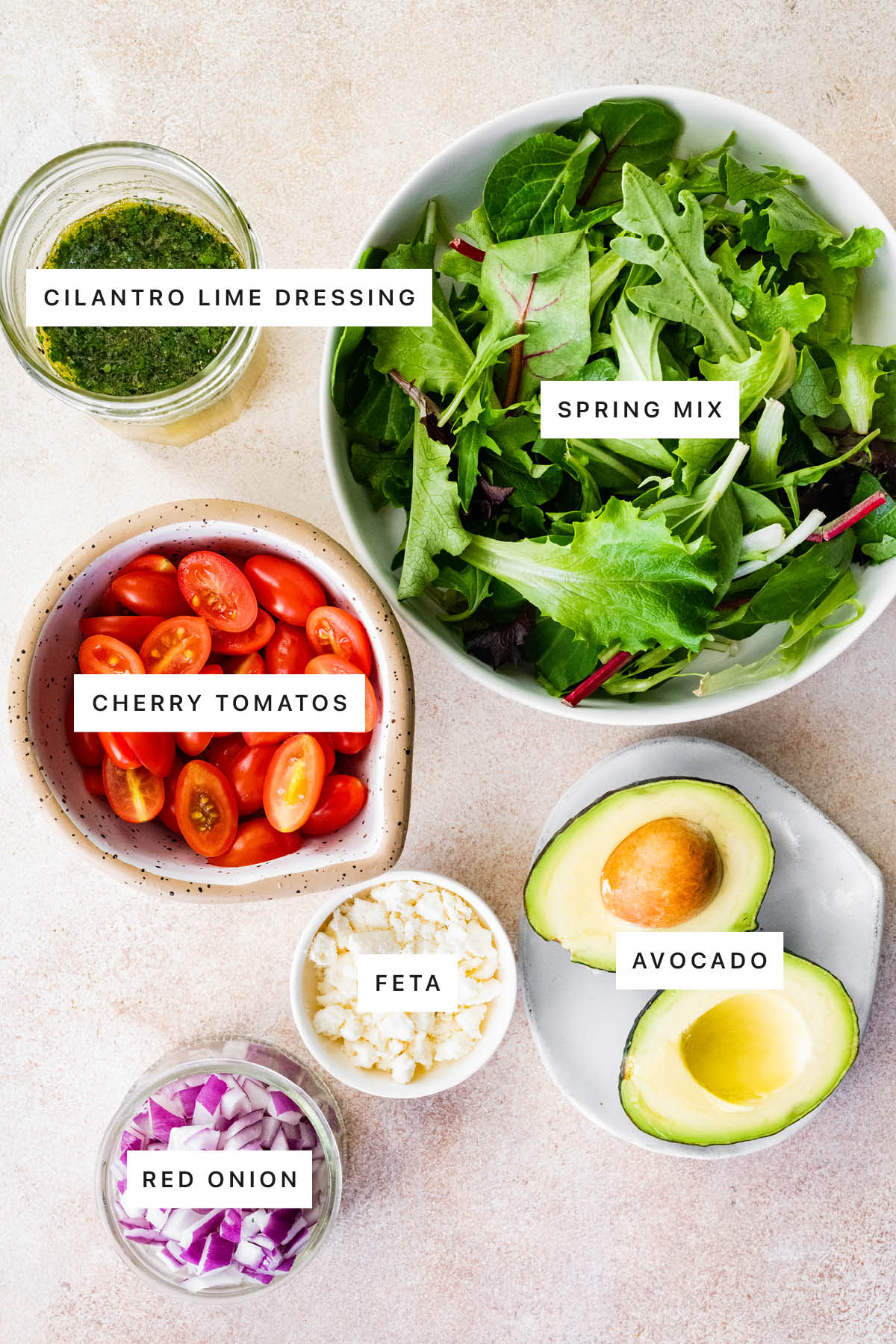 Ingredients measured out to make Deconstructed Guacamole Salad: cilantro lime dressing, spring mic, cherry tomatoes, feta, avocado and red onion.