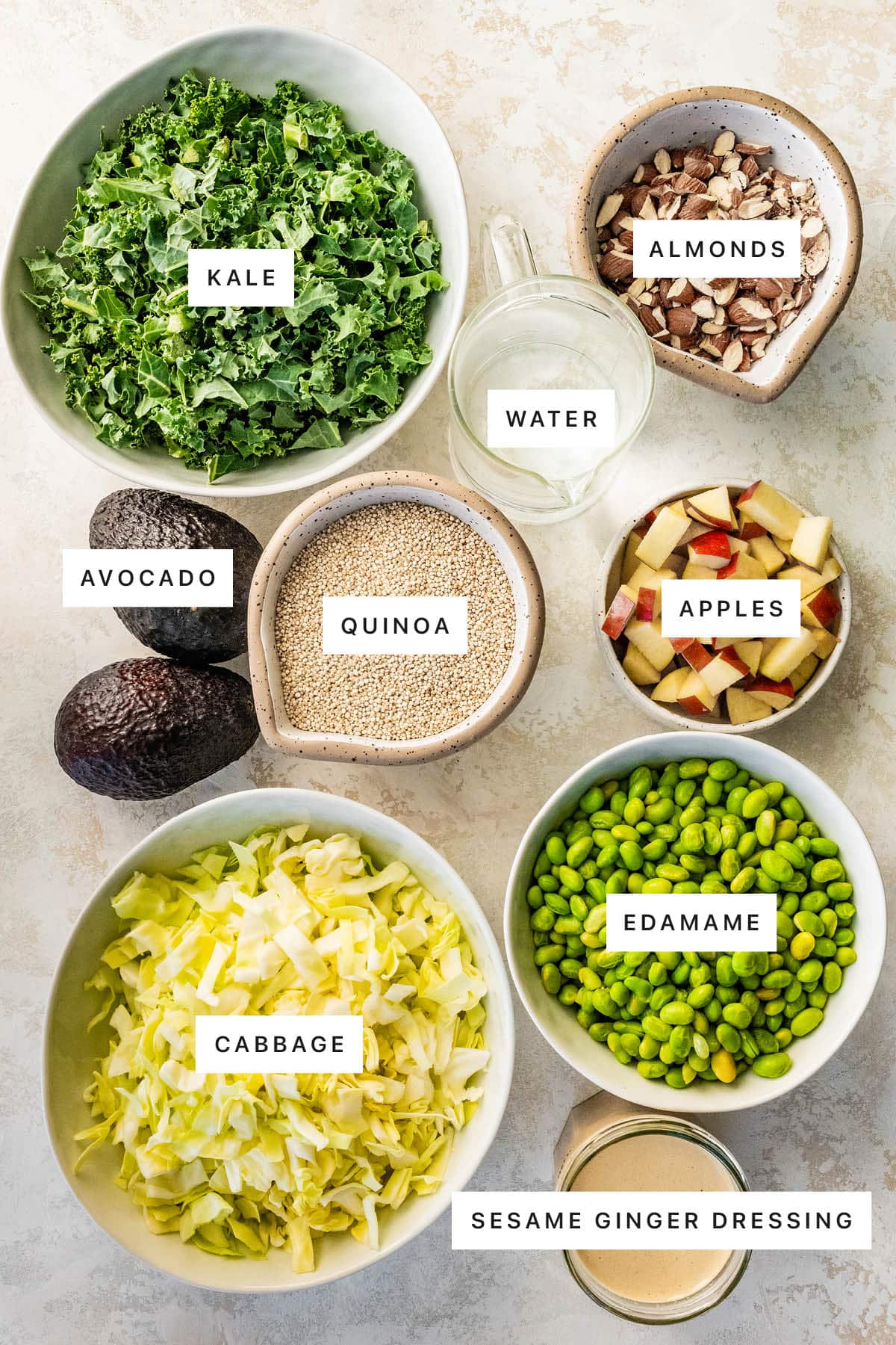 Ingredients measured out to make Meal Prep Edamame Salad: kale, almonds, water, quinoa, avocado, apples, cabbage, edamame and sesame ginger dressing.