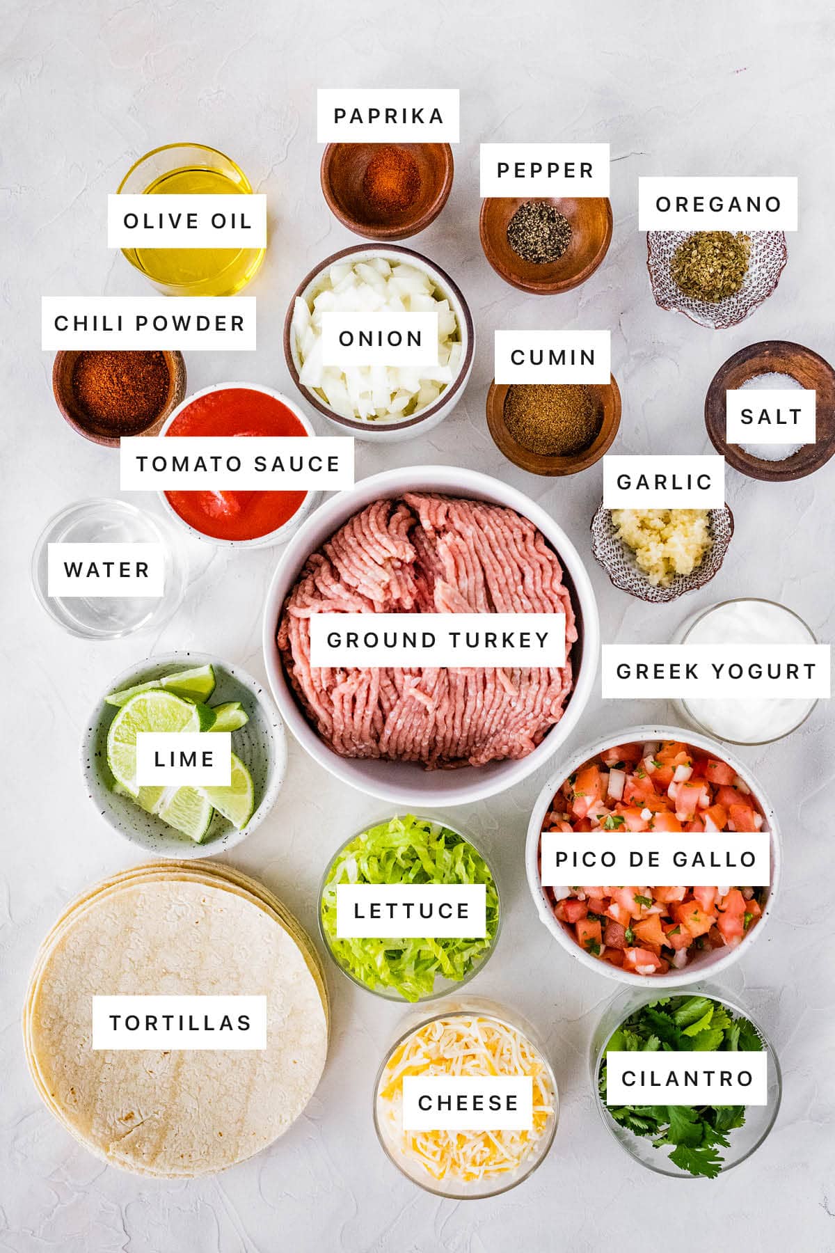 Ingredients measured out to make Ground Turkey Tacos: olive oil, paprika, pepper, oregano, chili powder, onion, cumin, salt, tomato sauce, garlic, water, ground turkey, Greek yogurt, lime, lettuce, pico de gallo, tortillas, cheese and cilantro.