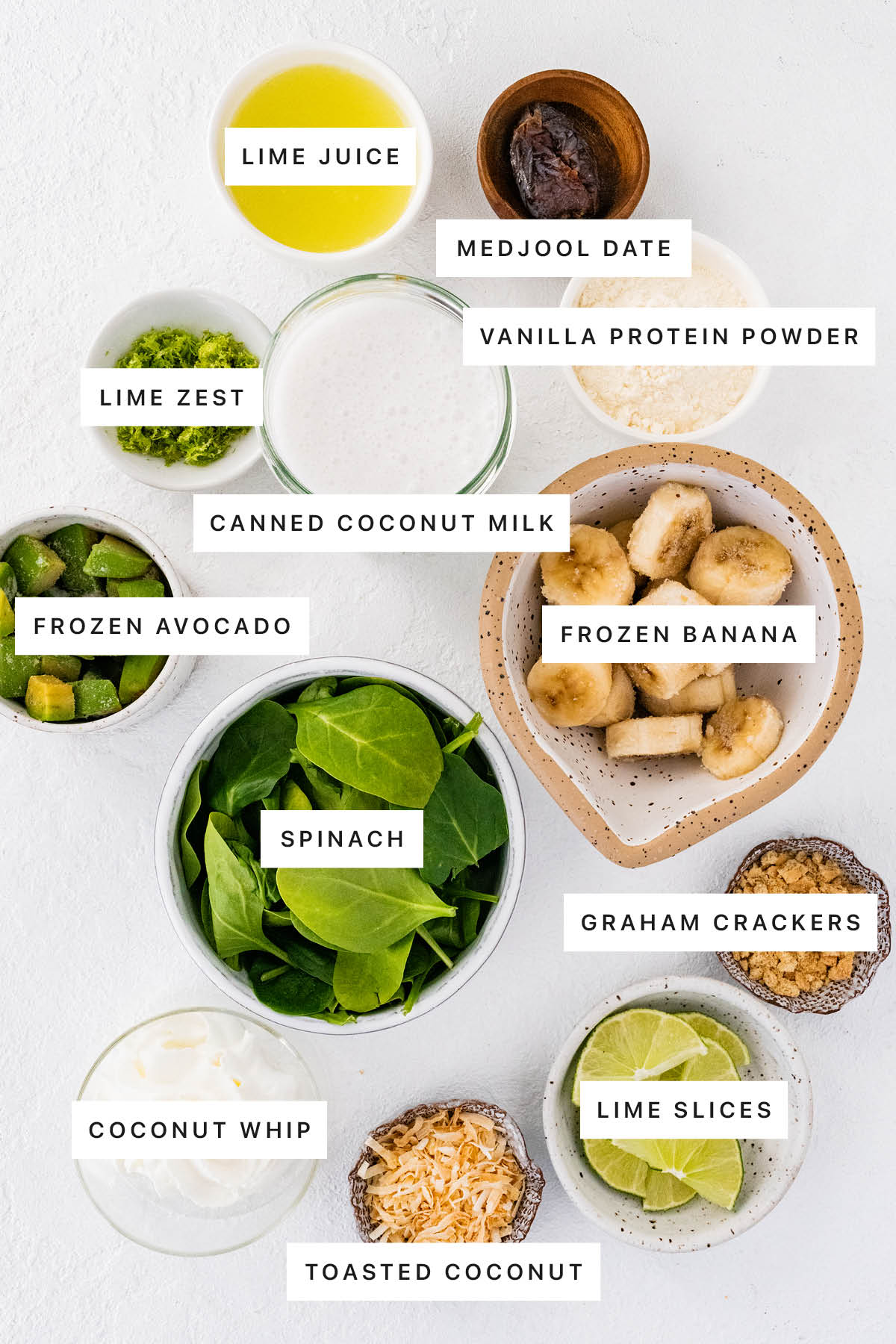 Ingredients measured out to make a Key Lime Pie Smoothie: lime juice, medjool date, vanilla protein powder, lime zest, canned coconut milk, frozen avocado, frozen banana, spinach, graham crackers, coconut whip, toasted coconut and lime slices.