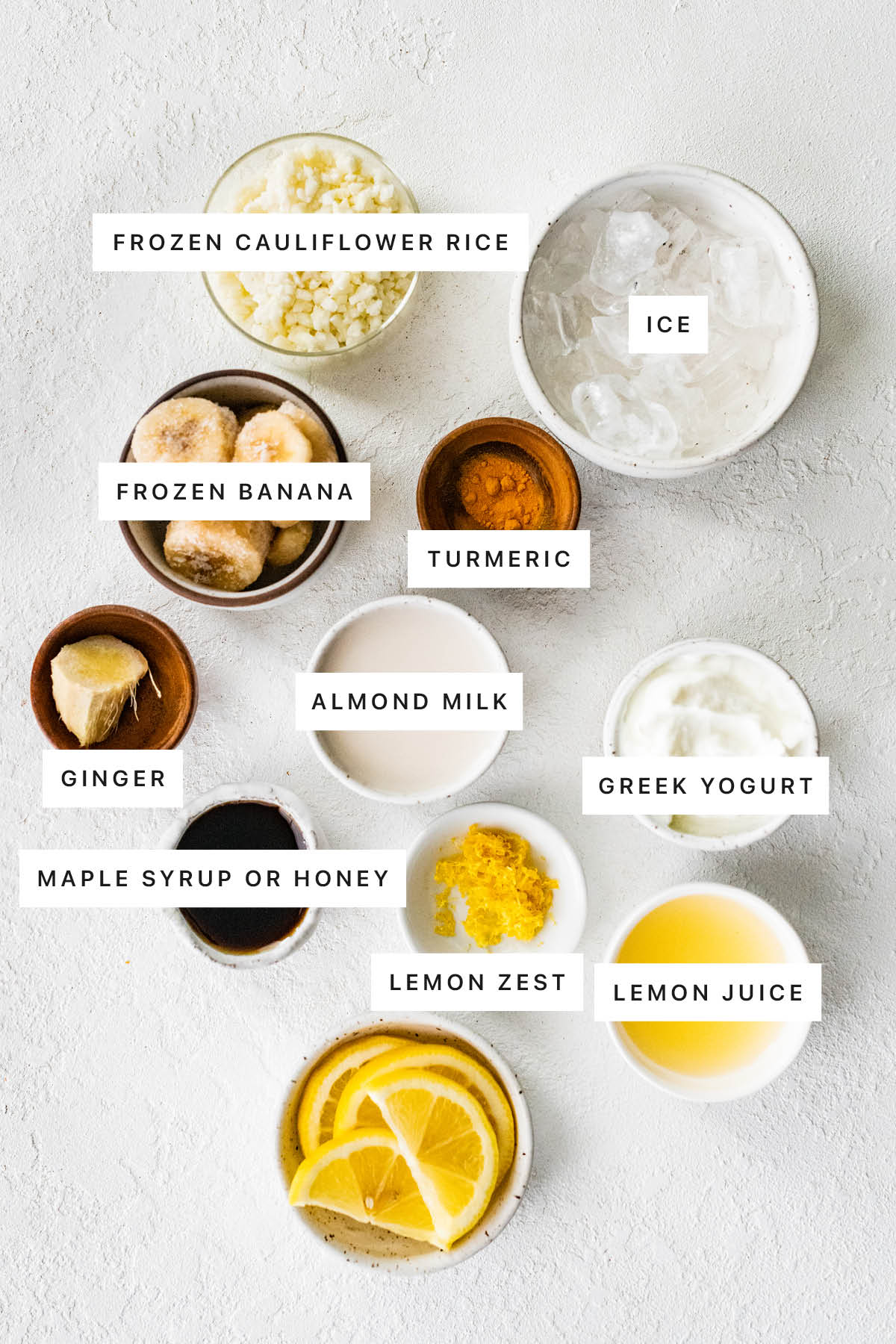 Measured ingredients to make a lemon smoothie: frozen cauliflower rice, ice, frozen banana, turmeric, ginger, almond milk, Greek yogurt, maple syrup, lemon zest, and lemon juice.