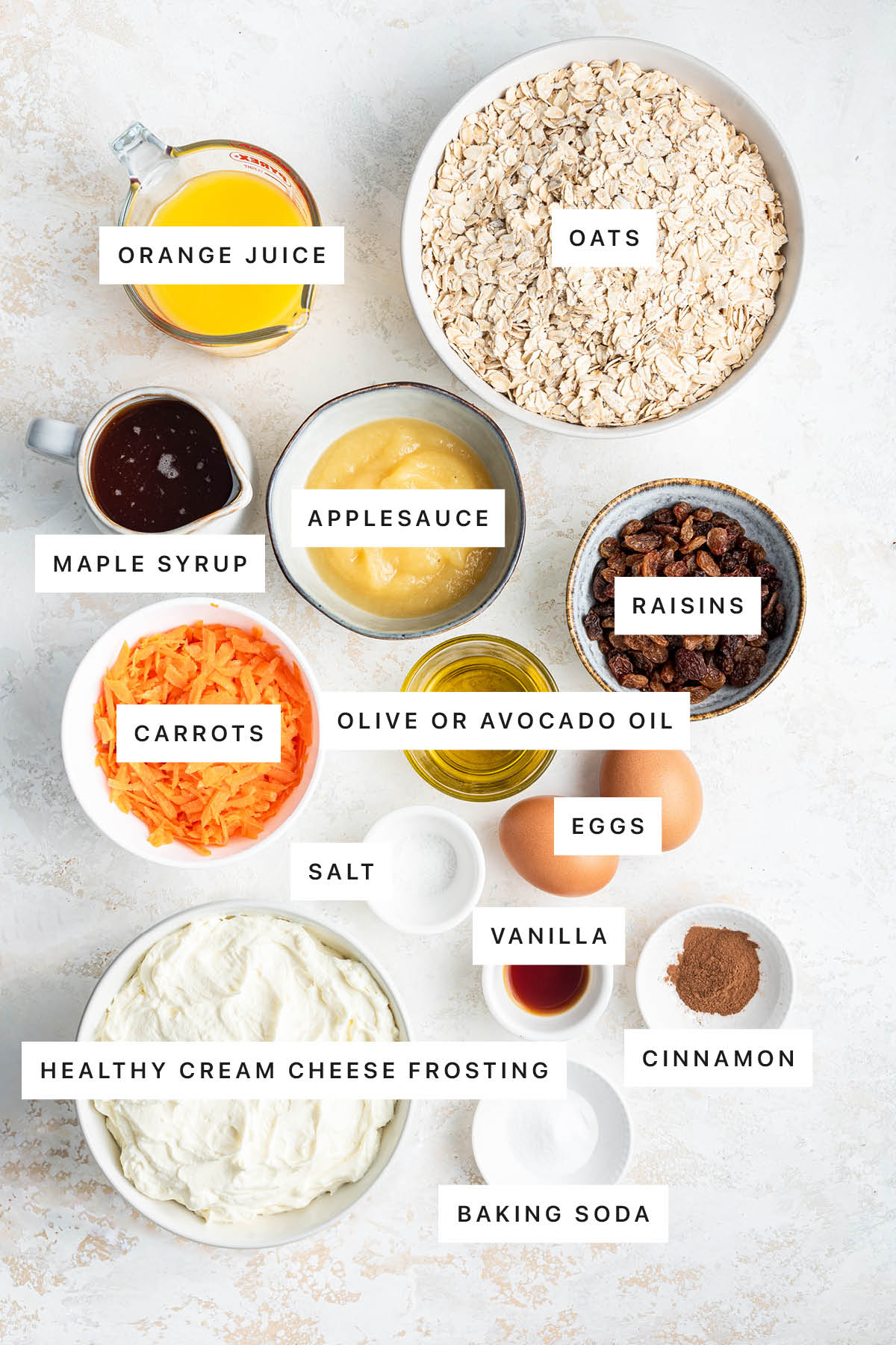 Ingredients measured out to make Oatmeal Carrot Cake: orange juice, oats, maple syrup, applesauce, raisins, carrots, olive oil, salt, eggs, vanilla, cinnamon, healthy cream cheese frosting and baking soda.