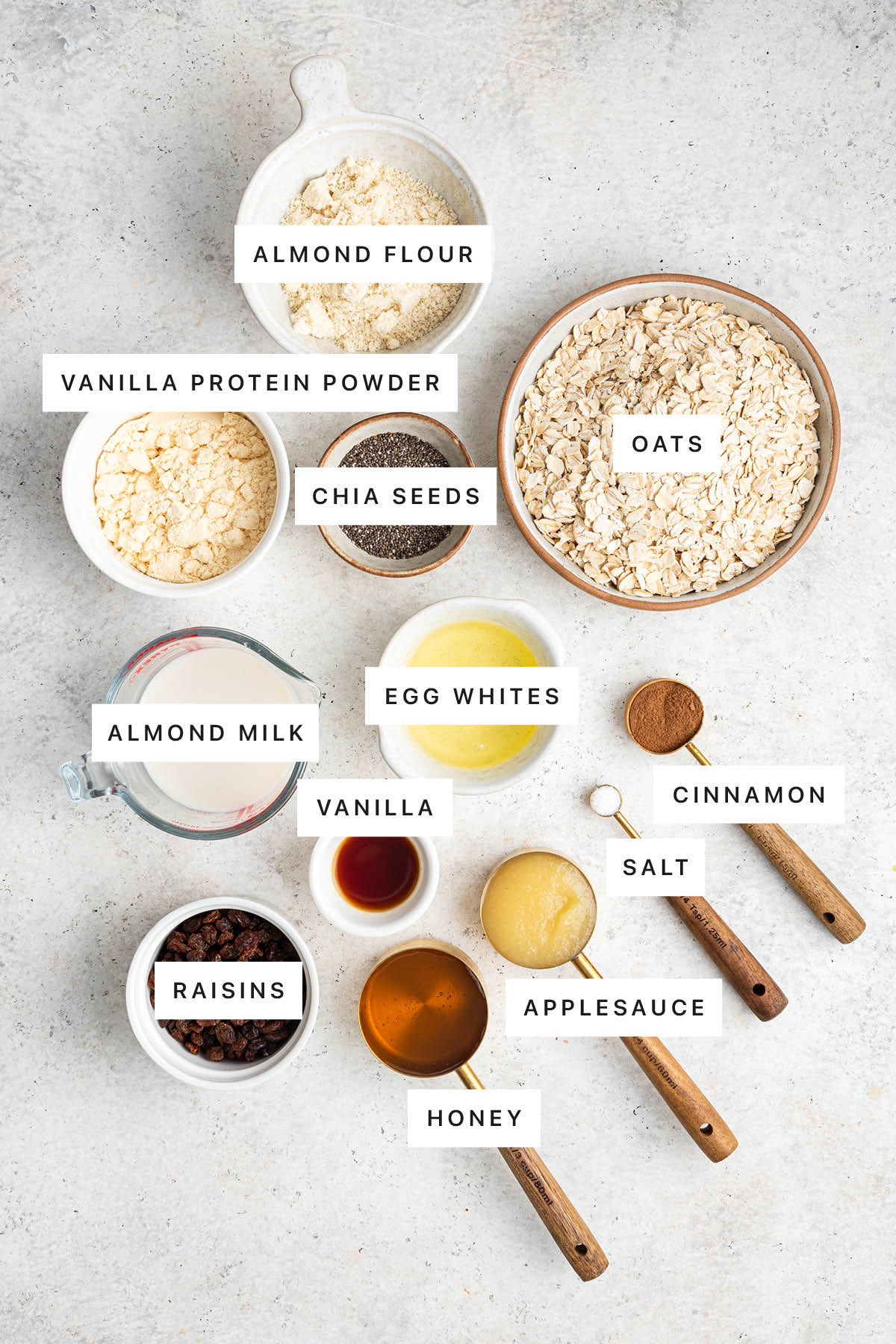 Ingredients measured out to make Oatmeal Raisin Protein Bars: almond flour, vanilla protein powder, chia seeds, oats, almond milk, egg whites, vanilla, raisins, honey, applesauce, salt and cinnamon.