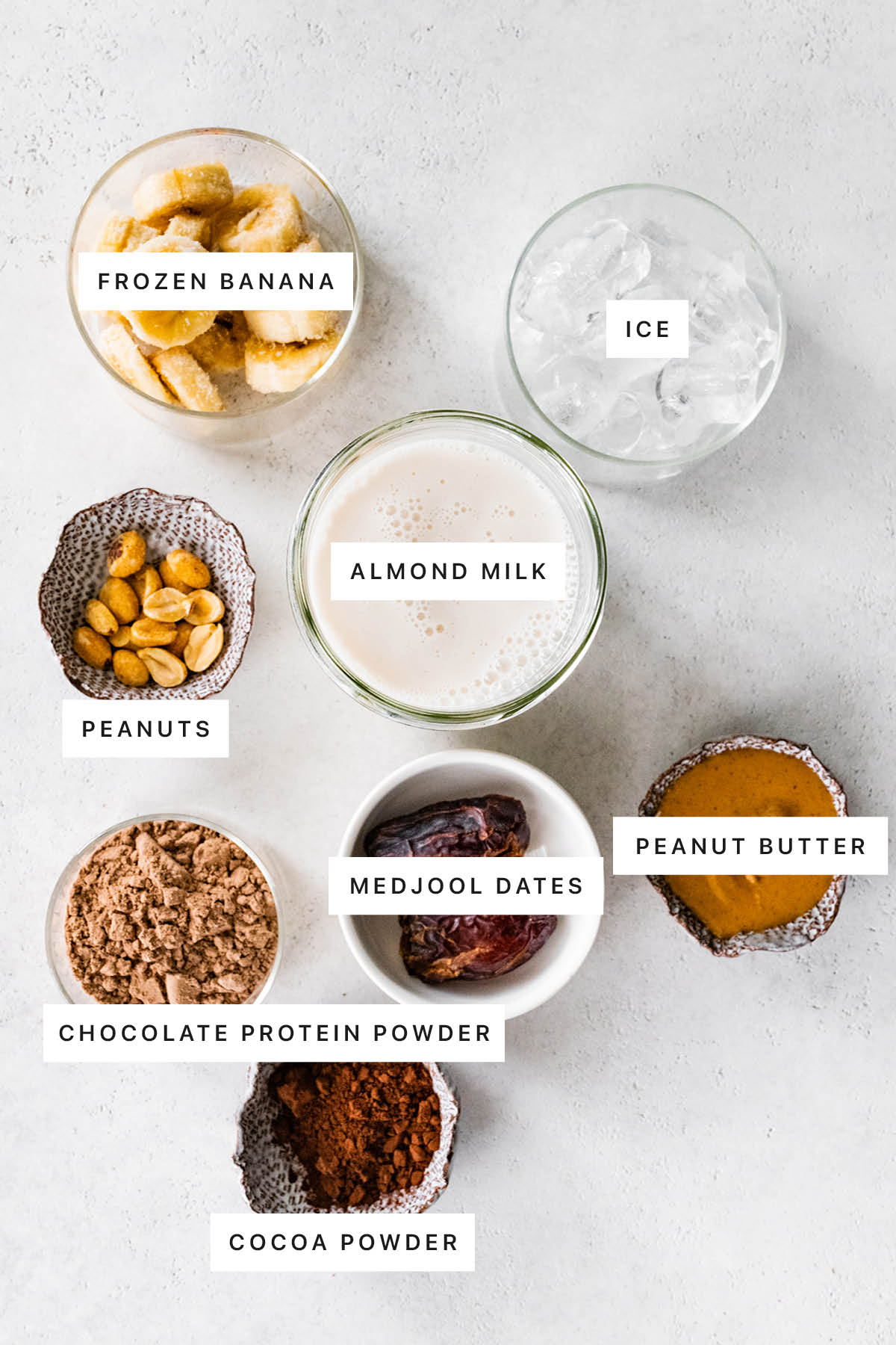 Ingredients measured out to make Snickers Protein Smoothie: frozen banana, ice, almond milk, peanuts, medjool dates, chocolate protein powder, peanut butter and cocoa powder.