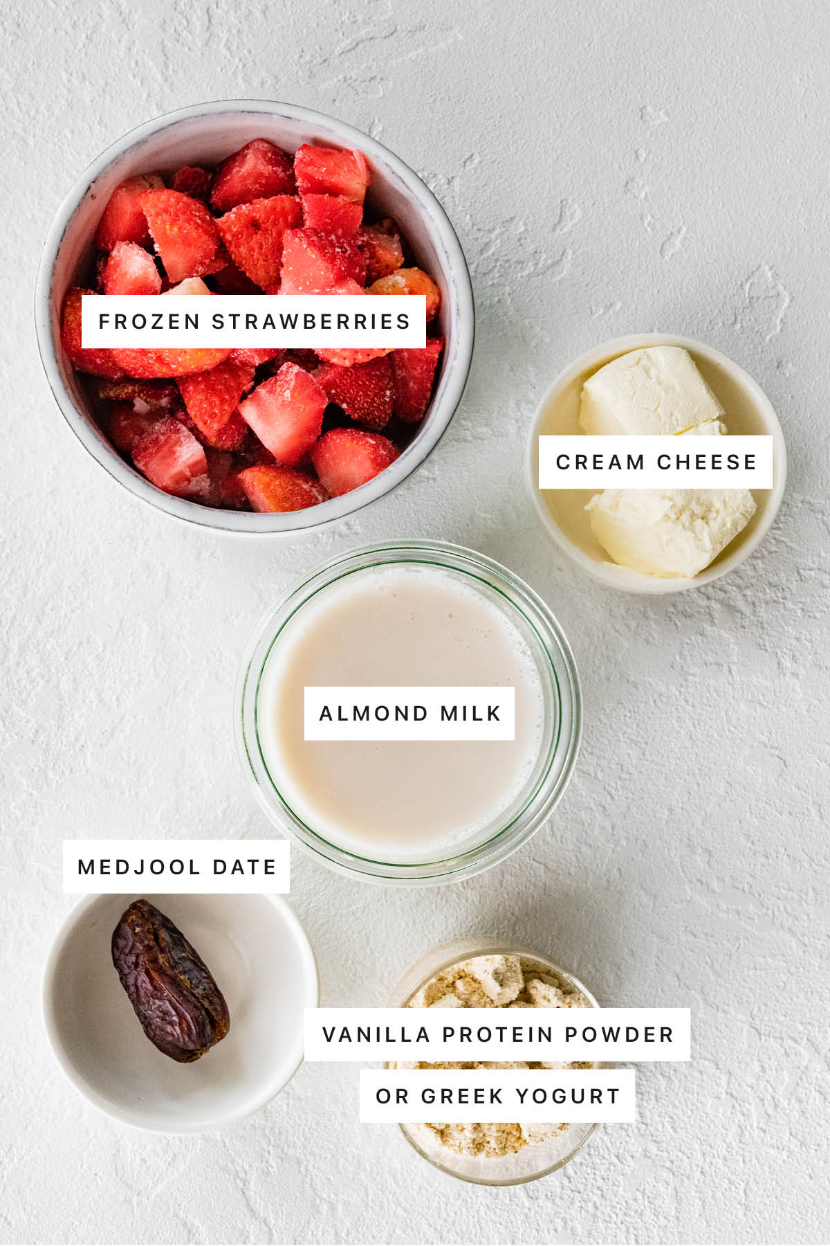 Measured ingredients to make a strawberry cheesecake shake: frozen strawberries, cream cheese, almond milk, medjool dates, and vanilla protein powder.