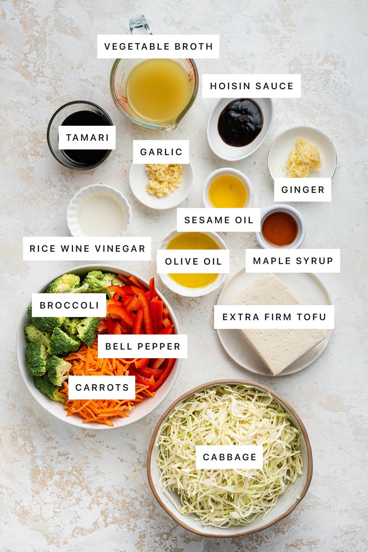 Ingredients measured out to make Tofu Cabbage Noodle Bowl: veggie broth, hoisin sauce, tamari, garlic, sesame oil, ginger, rice wine vinegar, olive oil, maple syrup, broccoli, tofu, bell pepper, carrots and cabbge.