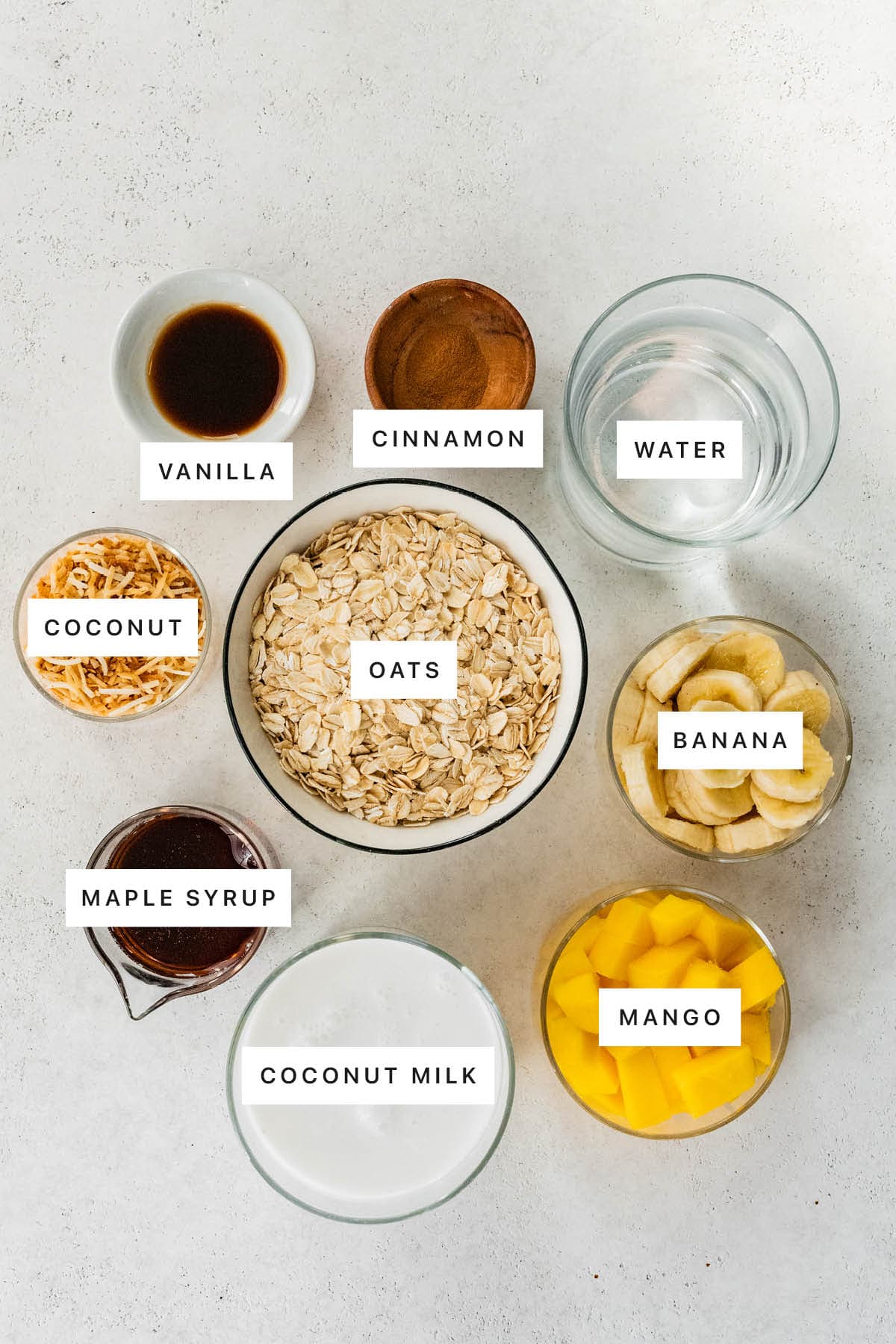 Ingredients measured out to make Tropical Oatmeal: vanilla, cinnamon, water, coconut, oats, banana, maple syrup, coconut milk and mango.