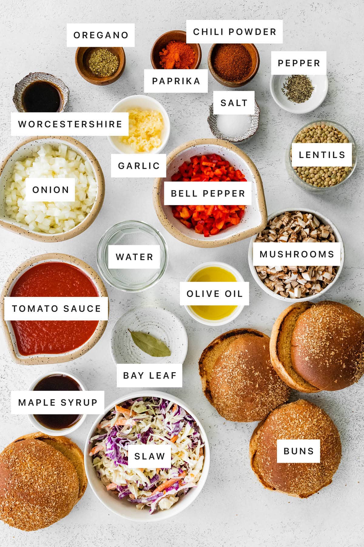 Ingredients measured out to make Vegan Sloppy Joes: oregano, paprika, chili powder, pepper, salt, Worcestershire, garlic, bell pepper, lentils, onion, water, olive oil, mushrooms, tomato sauce, bay leaf, maple syrup, slaw and buns.