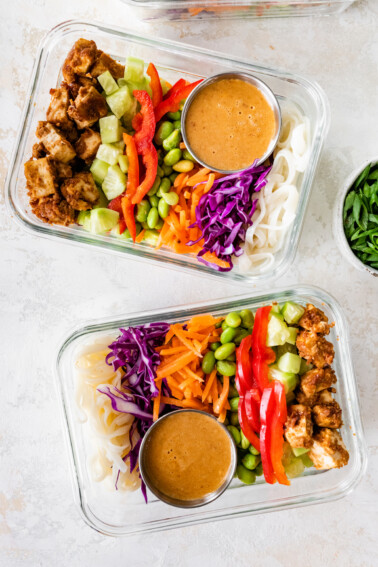 Two glass containers with the ingredients for the Asian noodle bowl including peanut baked tofu, red pepper, purple cabbage, edamame, rice noodles, celery, shredded carrots, and a sauce.