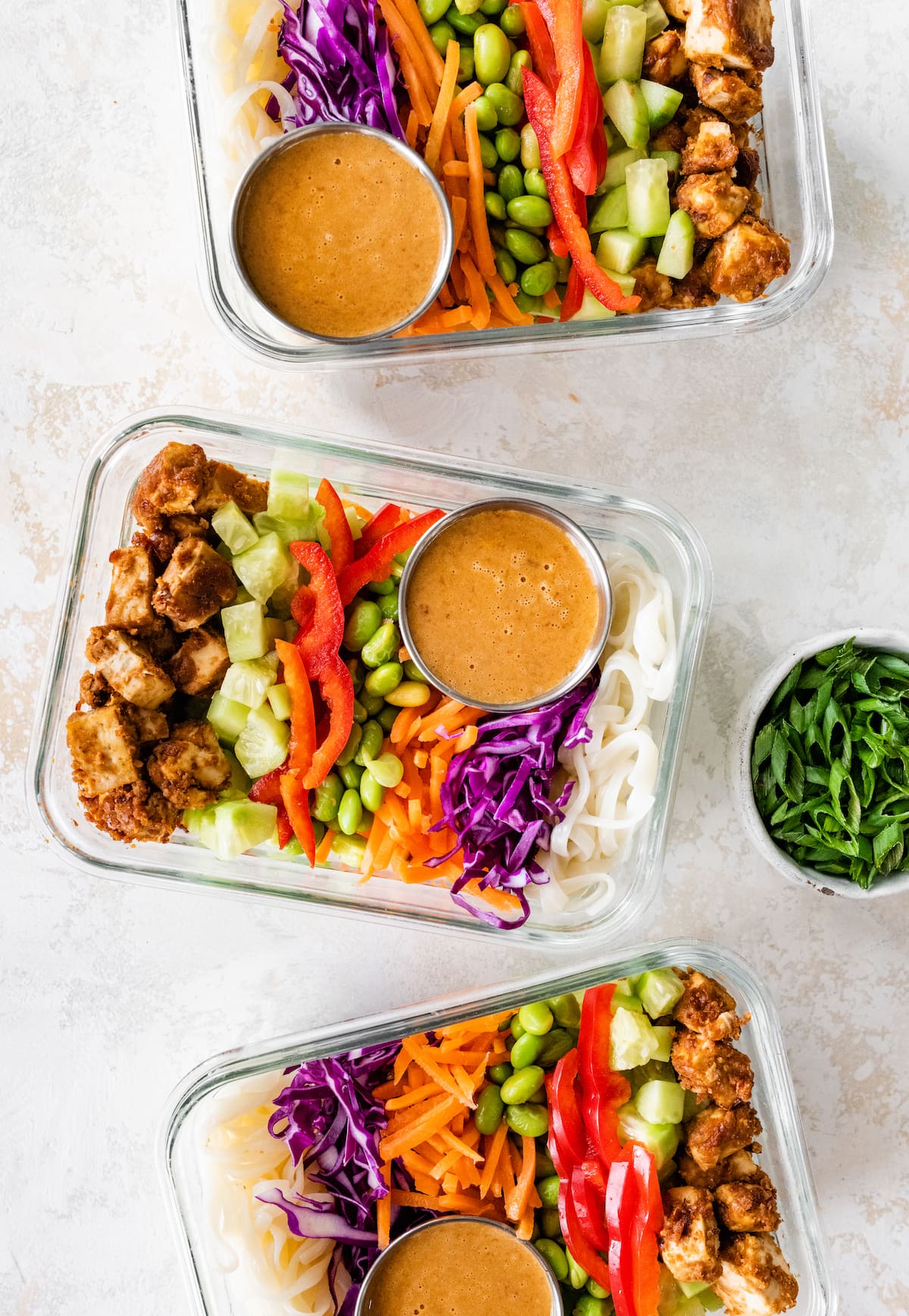 Three glass containers with the ingredients for the Asian noodle bowl including peanut baked tofu, red pepper, purple cabbage, edamame, rice noodles, celery, shredded carrots, and a sauce.