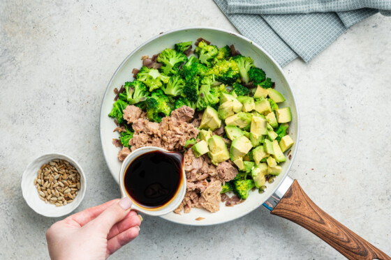 Woman's hand pouring sauce into skillet over tuna, broccoli, onions and avocado.