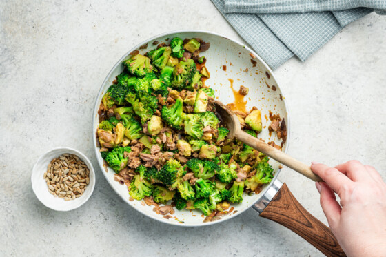 Woman's hand stirring broccoli avocado tuna bowl together in a skillet.