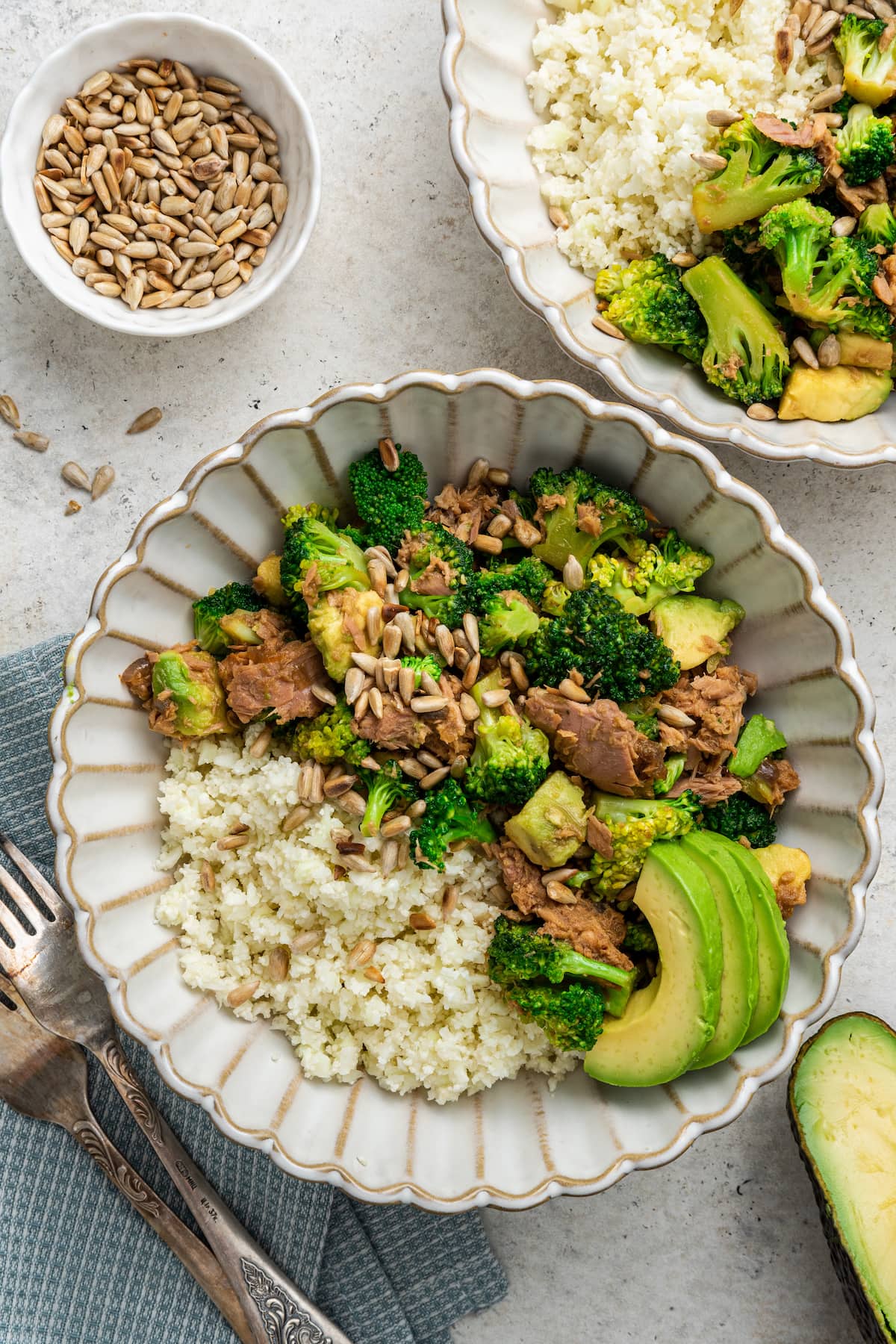 Two broccoli avocado tuna bowls topped with sunflower seeds and avocado slices and served with rice.